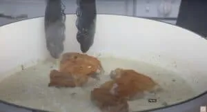 Crispy chicken breasts cooking in creamy garlic sauce, held by tongs in a white pan.